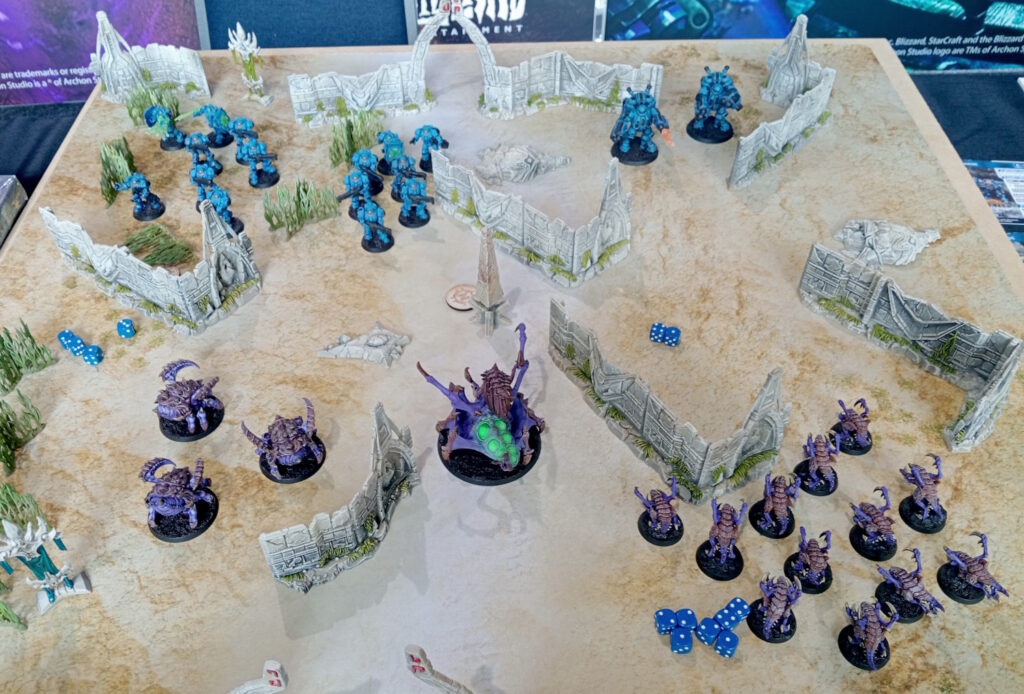 StarCraft TMG - laufende Partie Protoss gegen Zerg auf dem Demotisch mit Terrain und Wuerfeln (Quelle: https://www.tabletopbattles.com/first-look-the-starcraft-tabletop-miniatures-game)
